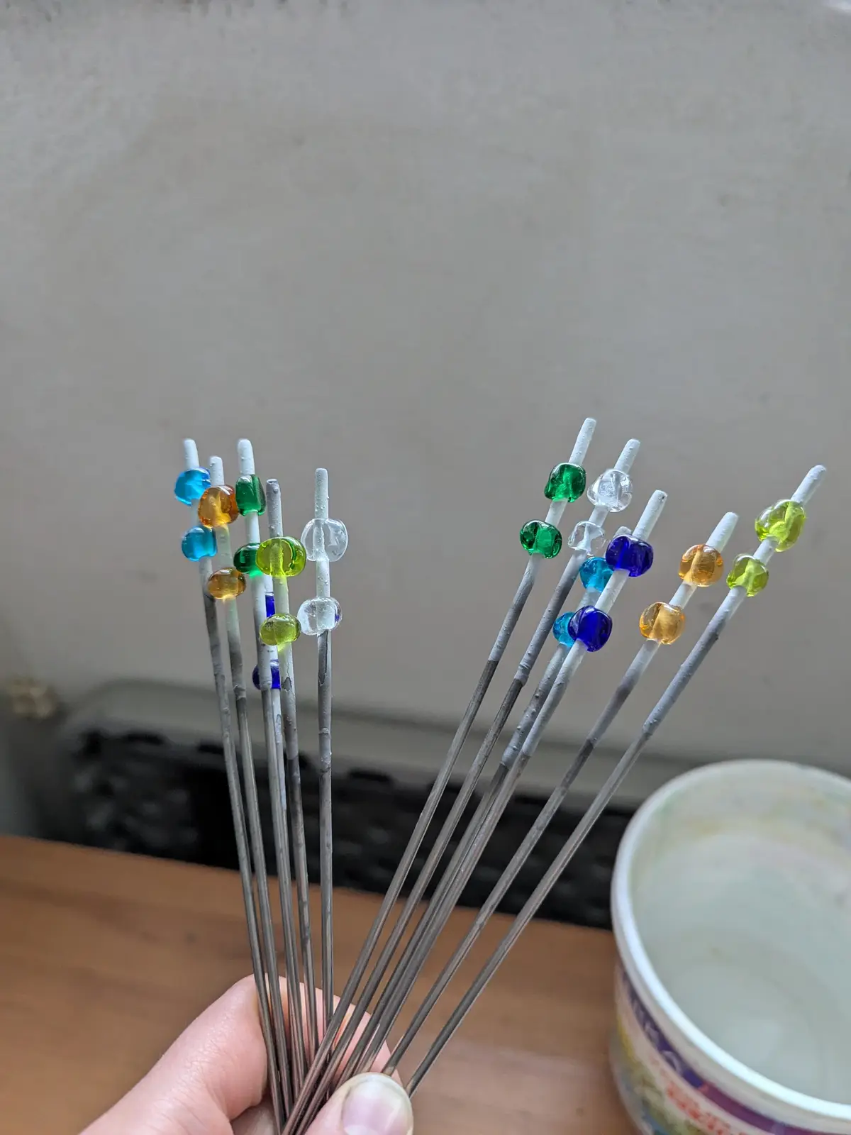 A hand holds multiple metal rods, each with a small, colorful glass bead fused near the top. The beads are various colors…