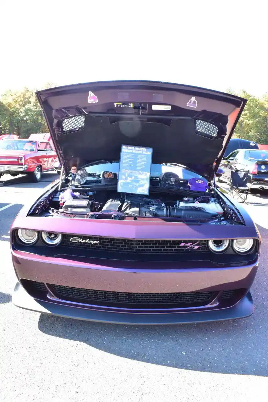 A purple Dodge Challenger R/T with its hood open at a car show, revealing a detailed engine bay. Inside the engine…