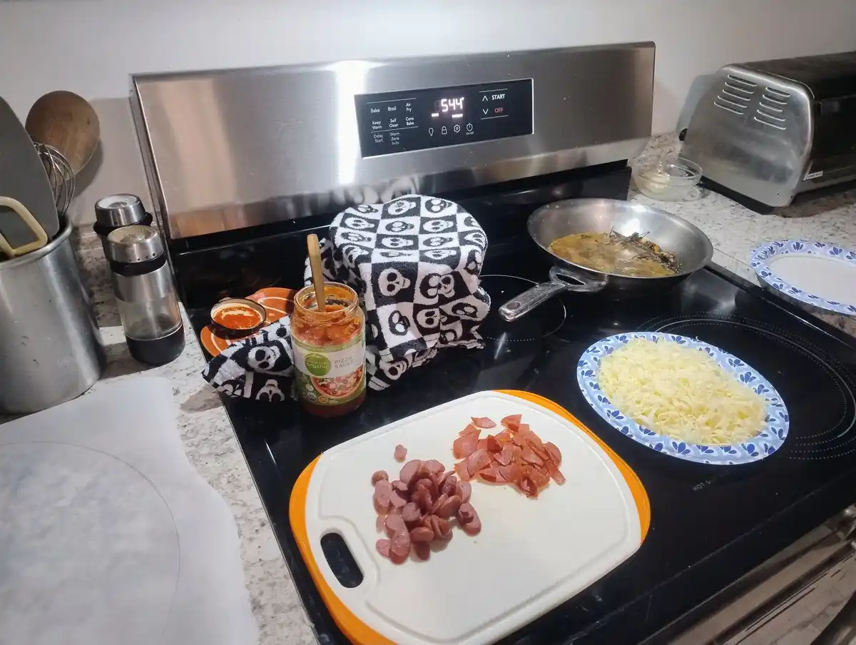 A kitchen stovetop is cluttered with pizza-making ingredients: a jar of pizza sauce, a plate of shredded cheese, and chopped…