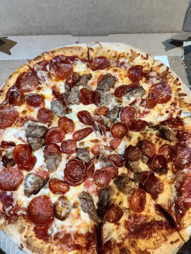 A close-up photo of a pepperoni and sausage pizza inside a cardboard box, with melted cheese and a golden-brown crust. The…