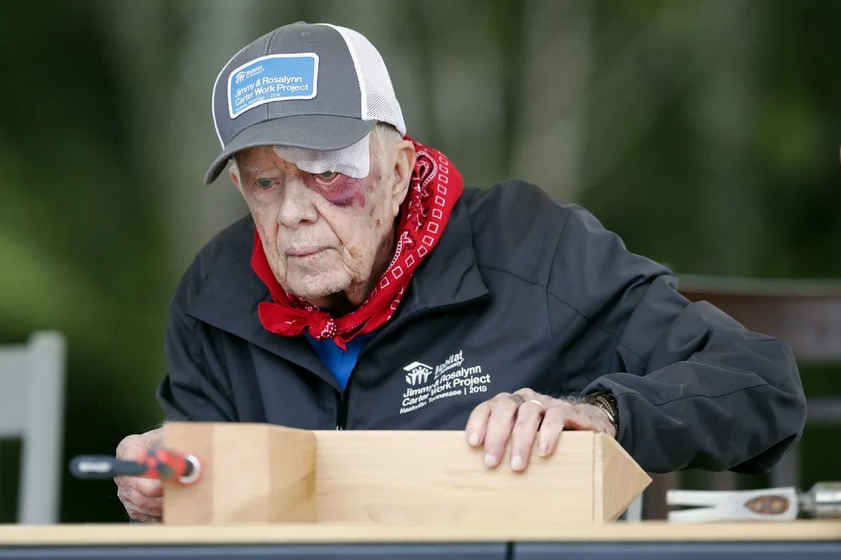 an elderly man with a bandaged eye and bruised face, wearing a 'Jimmy & Rosalynn Carter Work Project 2019' cap and jacket…