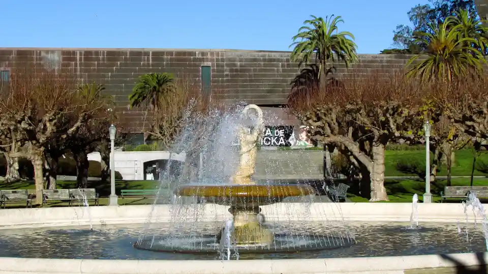 A photo of a large outdoor fountain with a central statue spraying water, set in front of a modern stone building with palm…