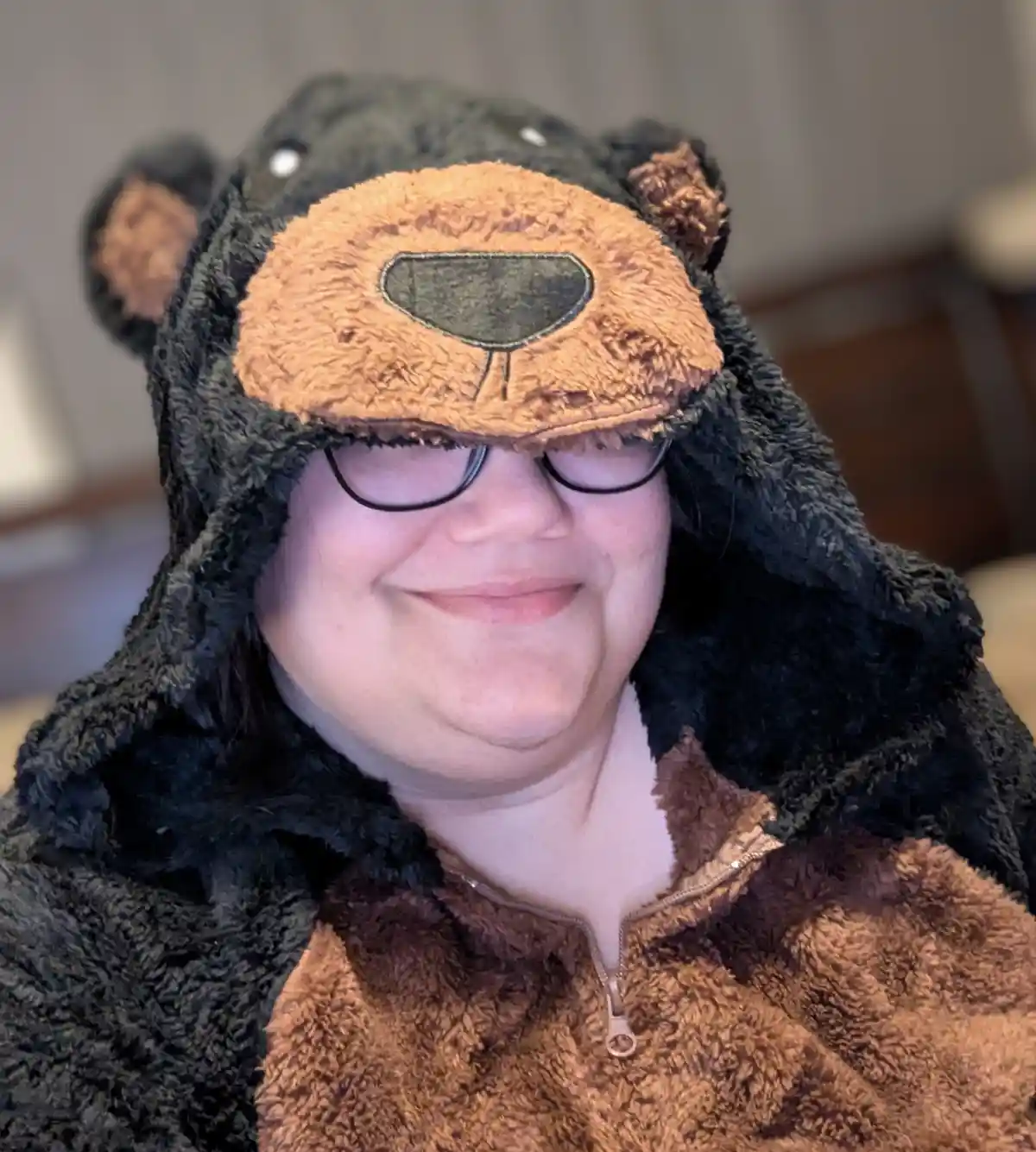 A person wearing a black and brown fuzzy bear onesie with a hood featuring a bear face, including ears and a nose, smiles…