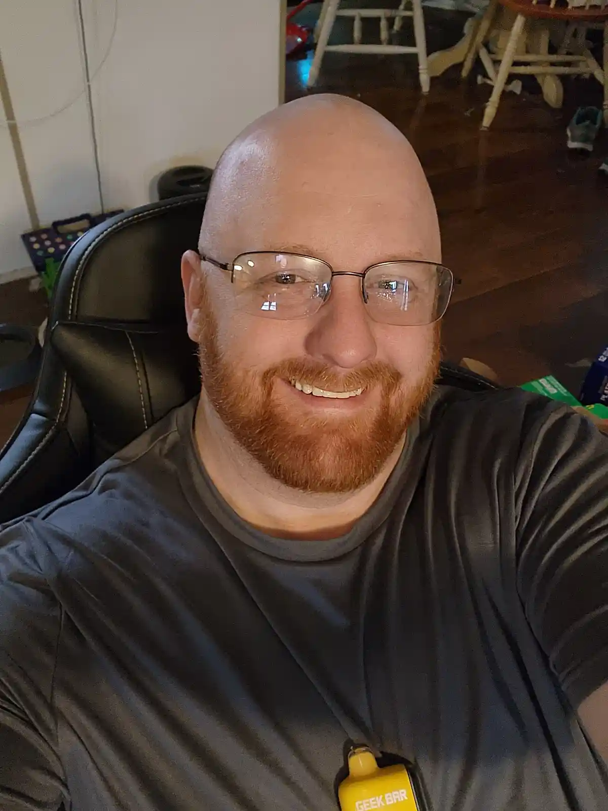 A bald man with a red beard and glasses smiles while sitting in a black gaming chair, wearing a dark grey t-shirt with a…