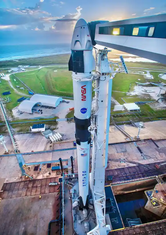 An aerial view of a SpaceX Falcon 9 rocket with a NASA logo and American flag, standing vertically on a launch pad next to a…
