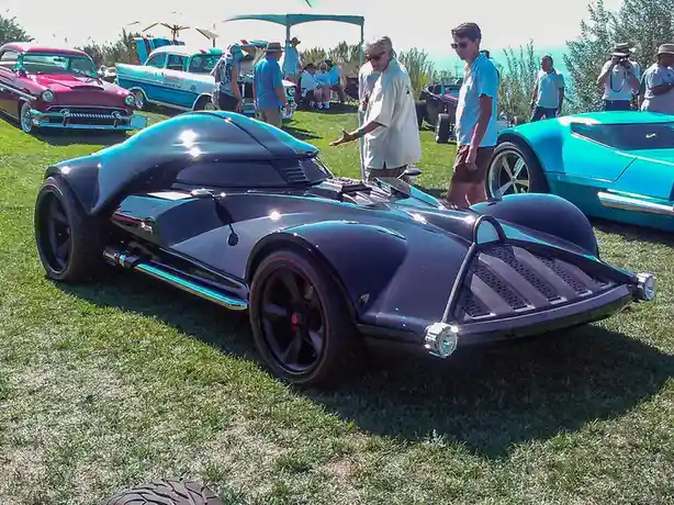 A shiny, custom-built black car with bat-like wings and a pointed front, resembling the batmobile from batman, is parked on…