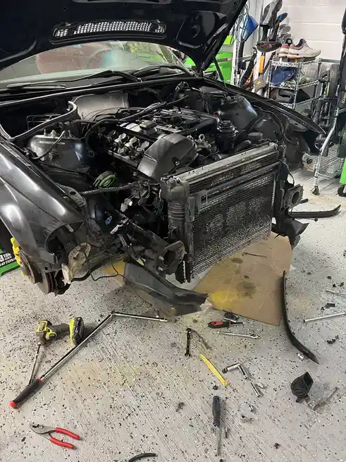 A car is shown in a garage with its hood open and engine bay exposed, the front bumper and radiator removed, revealing…