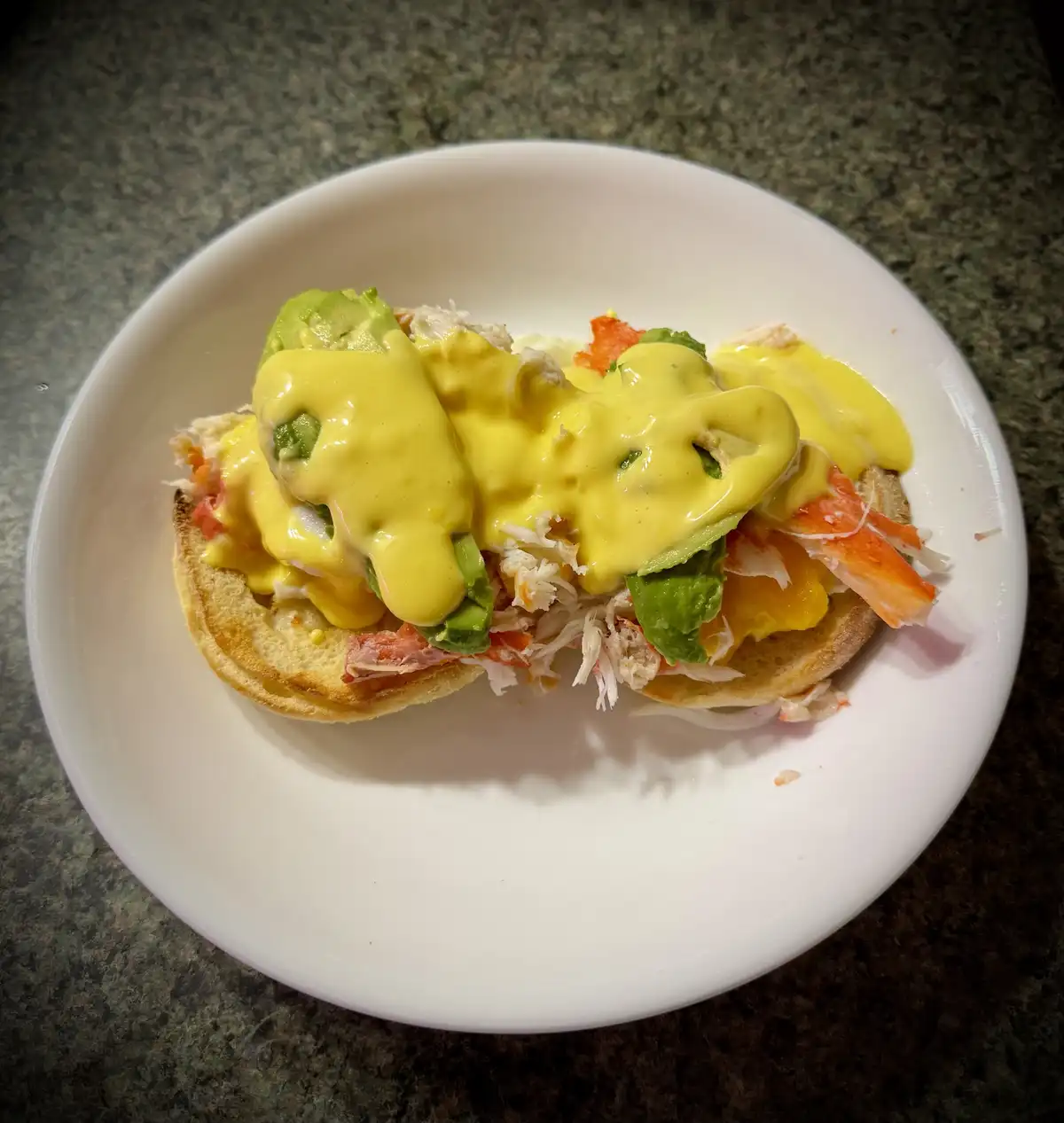 A close-up photo shows a white plate holding an open-faced sandwich on a toasted English muffin, topped with shredded crab…