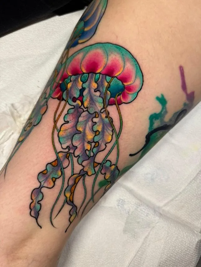 A close-up photo shows a colorful, detailed tattoo of a jellyfish on a person's forearm, featuring a pink and teal bell with…