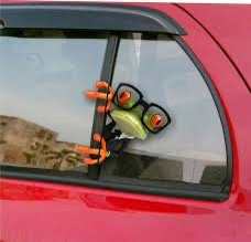 A cartoon frog wearing black-rimmed glasses and orange gloves is stuck to the inside of a red car's window, appearing to…