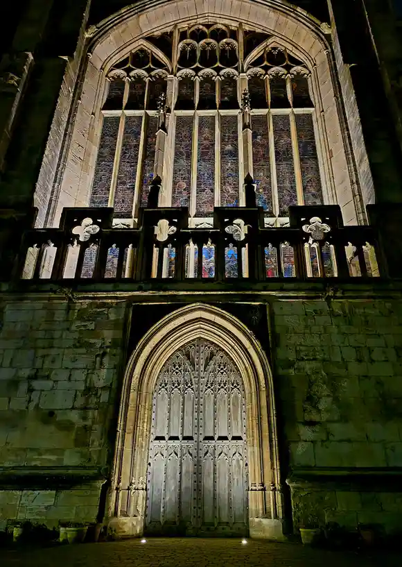 A nighttime photo of a grand Gothic church facade featuring an ornate wooden door and a large stained-glass window above it…