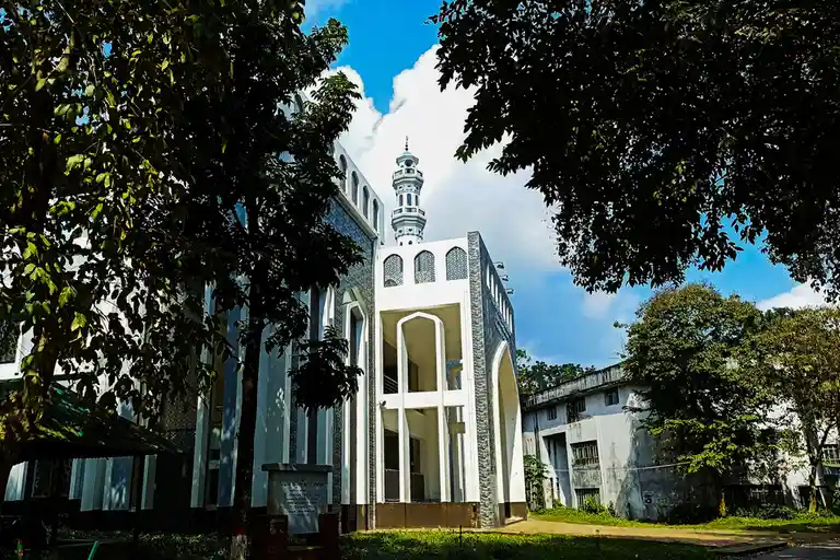 A photograph of a large, modern white mosque with a prominent minaret, framed by lush green trees against a blue sky with…