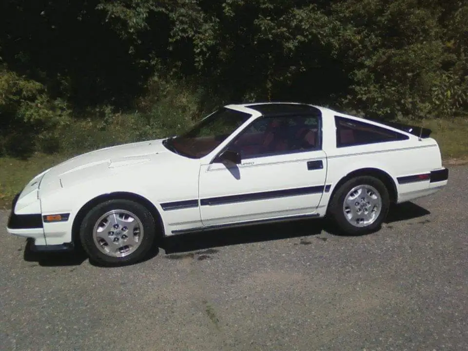 A white 1980s two-door sports car with black trim and a black roof is parked on asphalt with trees in the background. The…