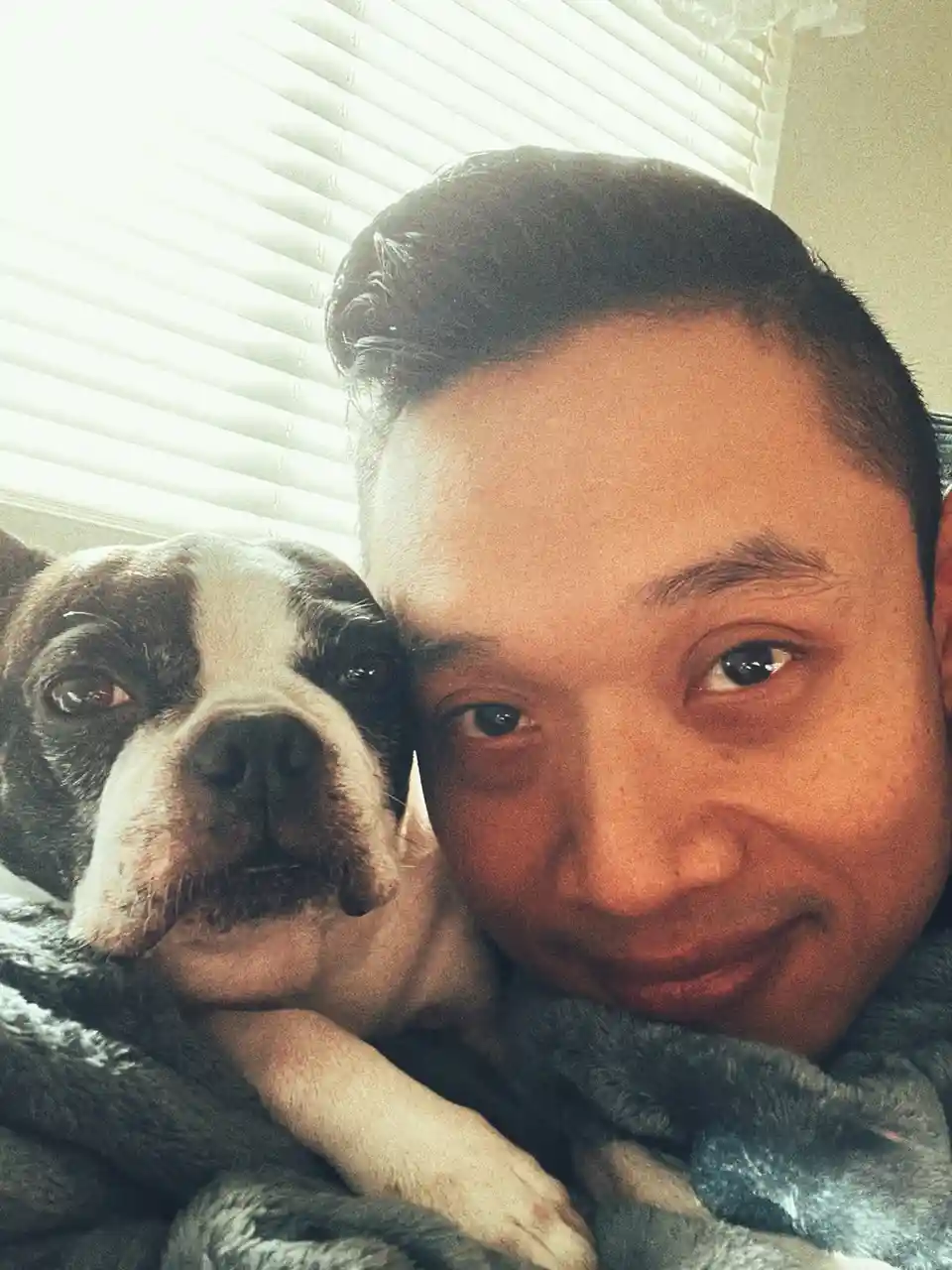 A close-up selfie shows a man with dark hair smiling at the camera while cuddling a black-and-white Boston Terrier dog. Both…