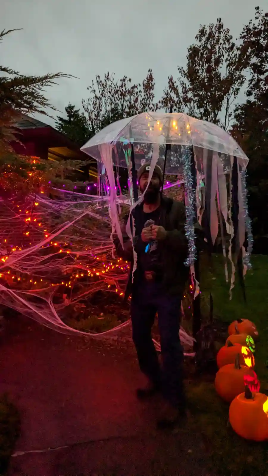 A man with a beard and beanie stands outdoors at dusk holding a clear umbrella decorated with string lights and hanging…