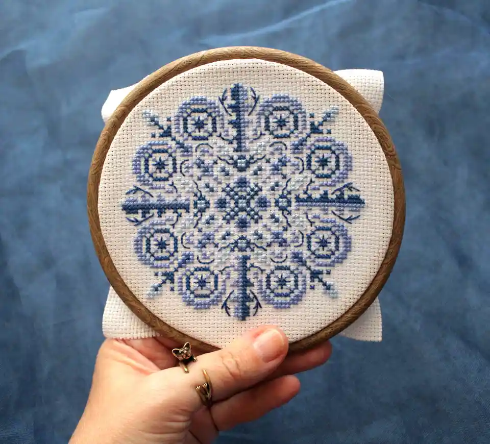 A hand holds a wooden embroidery hoop displaying a cross-stitch pattern stitched in blue thread on white fabric, forming the…