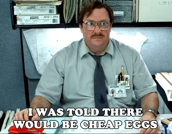 A man with glasses and a mustache sits at a cluttered office desk, wearing a striped shirt and tie with a name badge. He has…