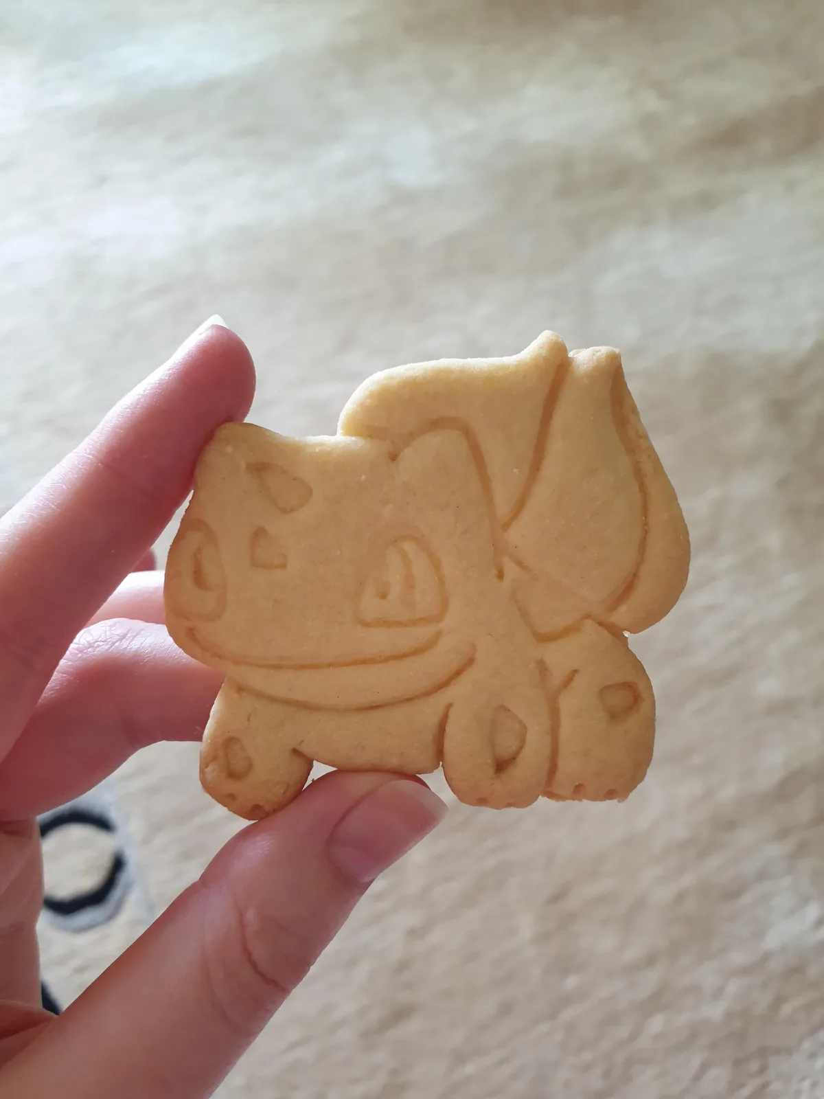 A hand holds up a plain, uniced, golden-brown sugar cookie shaped like the Pokémon Bulbasaur, with simple embossed lines…