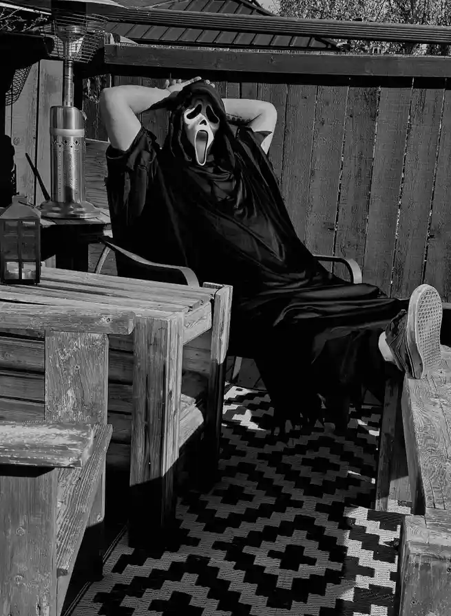 A person wearing a black hooded robe and the iconic white Ghostface mask from the 'Scream' movie franchise is lounging in a…