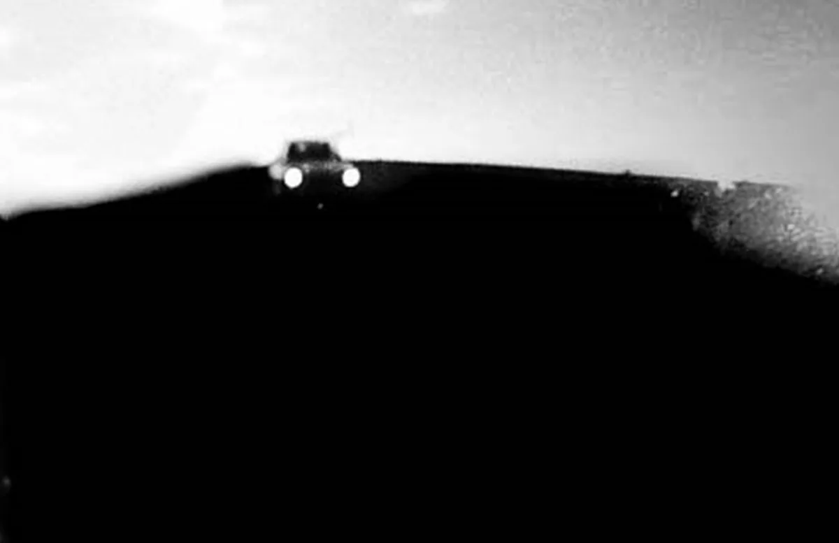 A grainy black-and-white photo shows a car's headlights illuminating the top of a dark hill or ridge at night. The rest of…