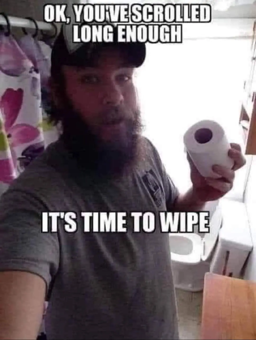 A man with a beard and cap stands in a bathroom holding a roll of toilet paper, looking directly at the camera. Text…