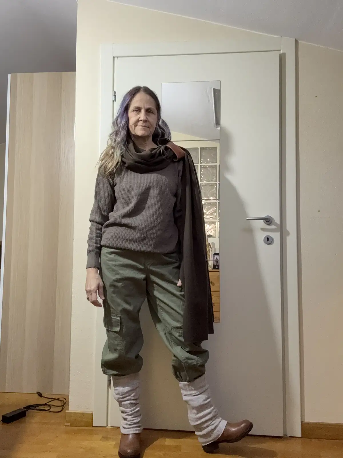 A woman with purple and blonde hair stands in front of a white door with a mirror inset, wearing a grey sweater, olive green…