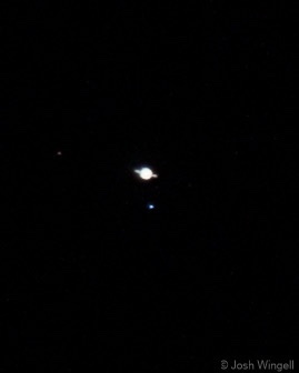 A blurry, low-resolution photo of Saturn in the night sky, appearing as a small, bright, slightly off-white disc with faint…