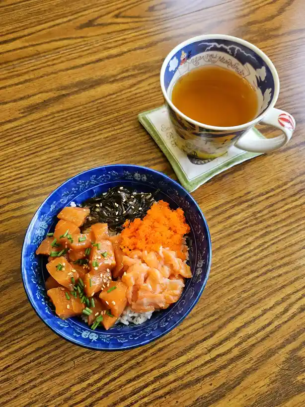A top-down photo shows a blue bowl of salmon donburi with rice, salmon chunks, seaweed, and orange roe on a wooden table…