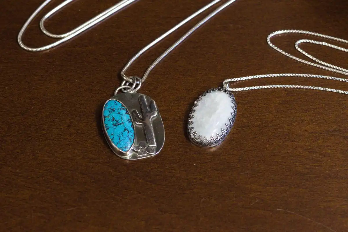 Two silver necklaces with oval pendants lie on a dark wooden surface. The left pendant features a bright blue turquoise…