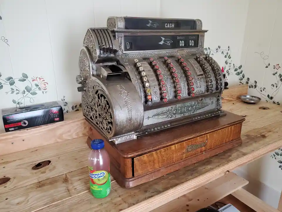 An ornate, vintage National brand cash register sits on a wooden counter next to a small bottle of Snapple kiwi-strawberry…
