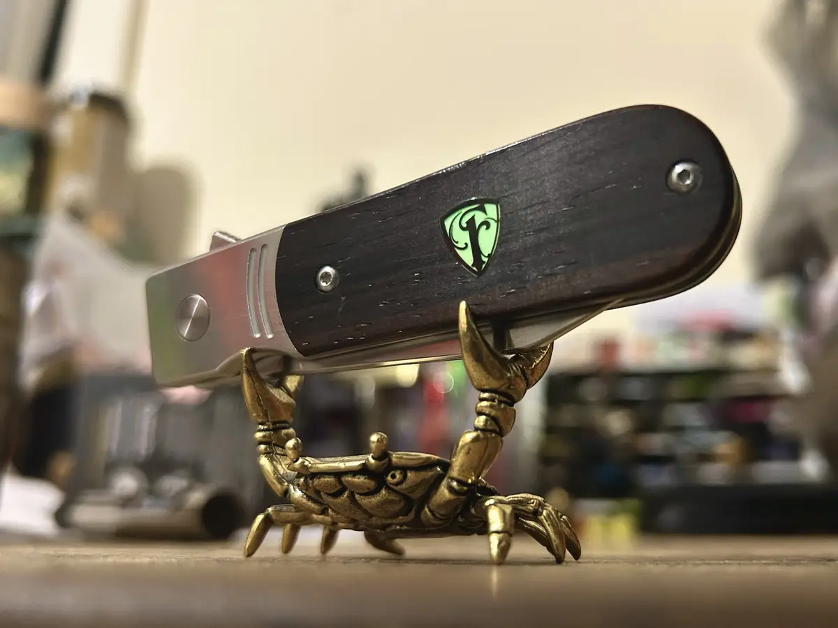 A close-up photo shows a brass crab figurine holding up a folding knife with a dark wooden handle and a glowing green logo…