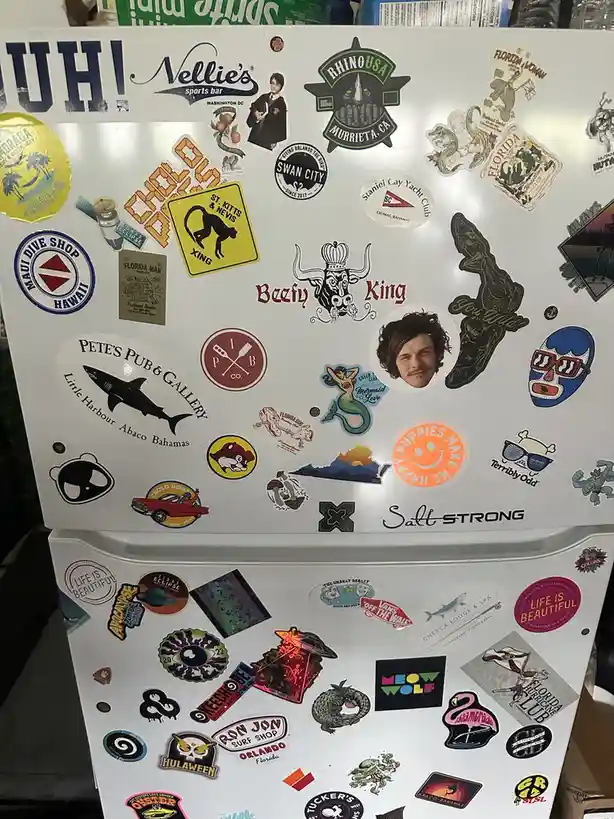 A white refrigerator door is covered in dozens of colorful, overlapping stickers from various places like Florida, Hawaii…