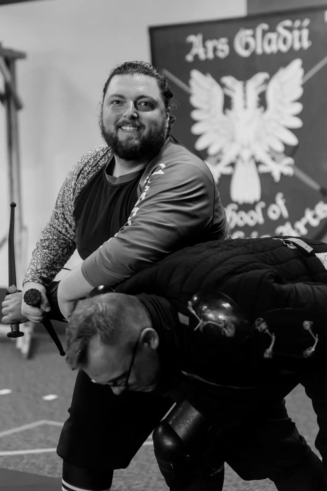 A black-and-white photo shows a man in a martial arts studio holding a sword while pinning another man in protective gear…