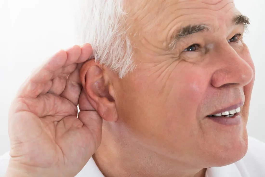 a close-up of an older man with white hair smiling while cupping his hand behind his ear, as if straining to hear something…