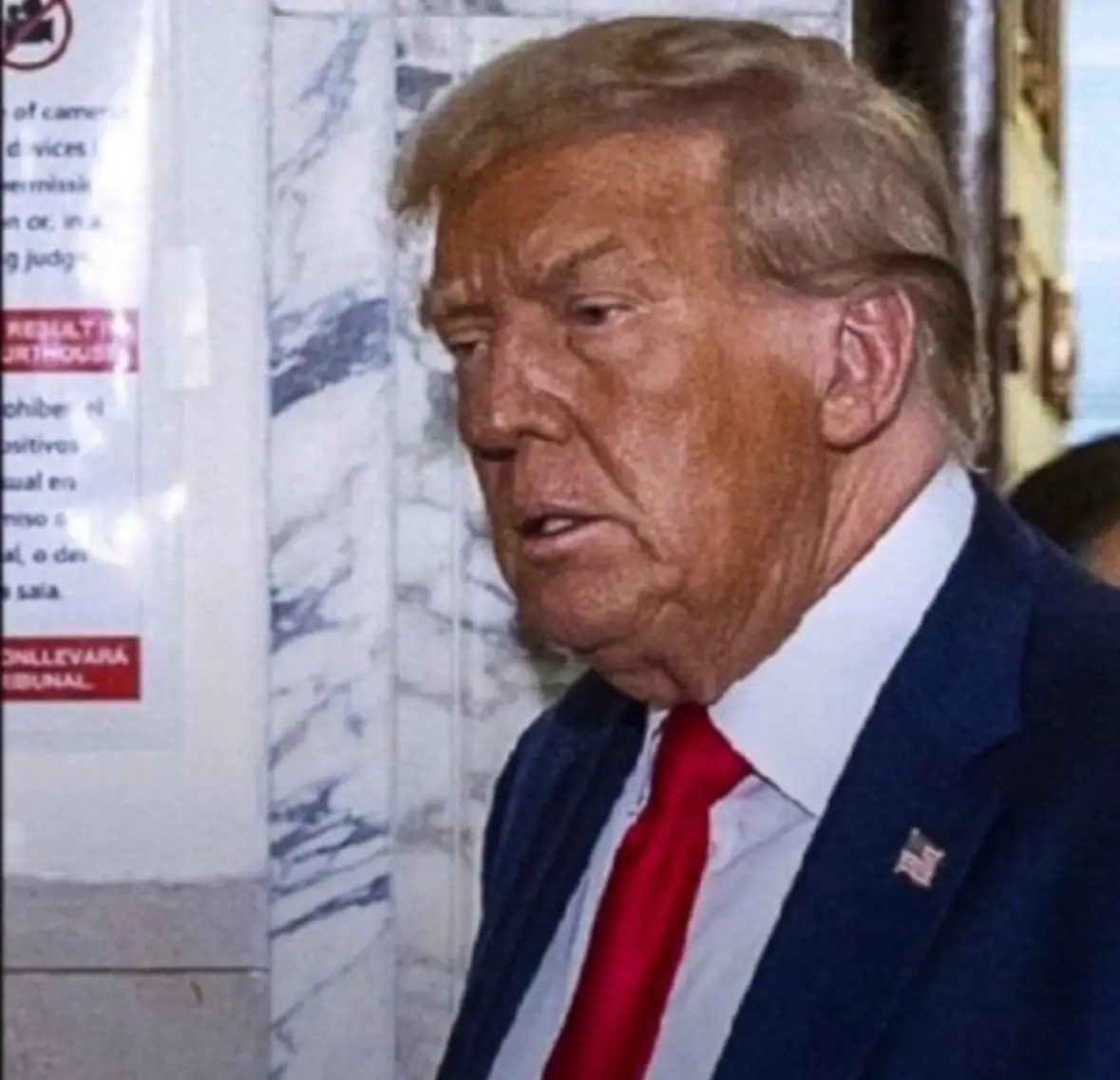 A man with blond hair, wearing a dark suit, white shirt, and red tie, looks slightly to his left with a confused or…