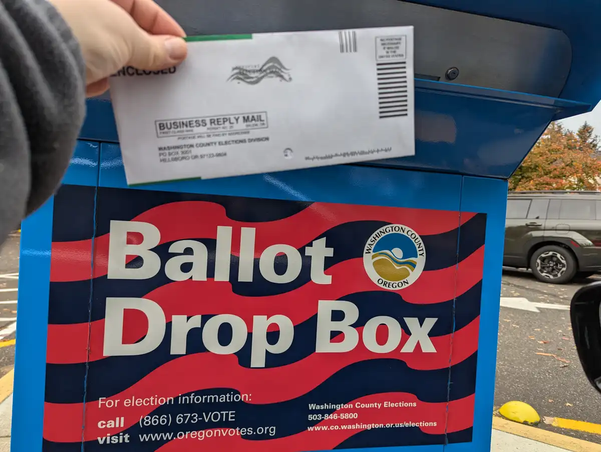 A hand holds a white business reply mail envelope above a blue ballot drop box labeled 'Ballot Drop Box' for Washington…