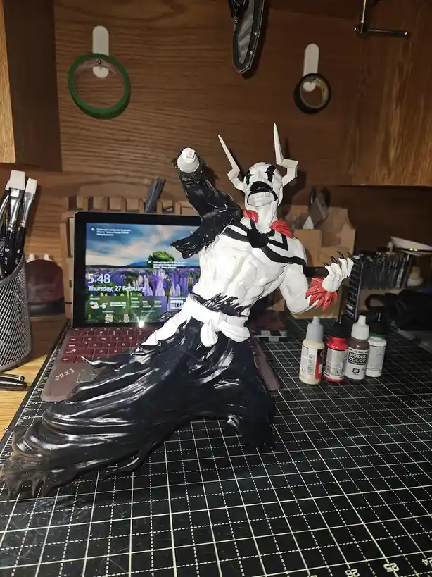 A painted anime-style figurine of a muscular, white-skinned character with black markings, horns, and red accents is posed…