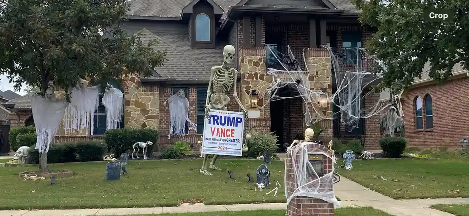 A house decorated for Halloween with numerous skeletons, ghosts, and spiderwebs. A large skeleton in the front yard holds a…