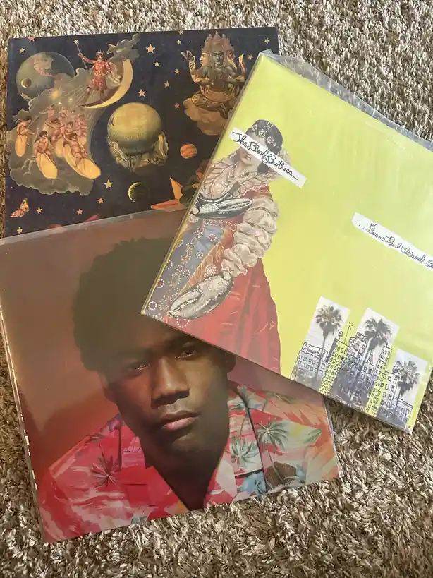 Three vinyl record covers are stacked on a beige carpet. The top cover features a surreal, cosmic scene with planets…