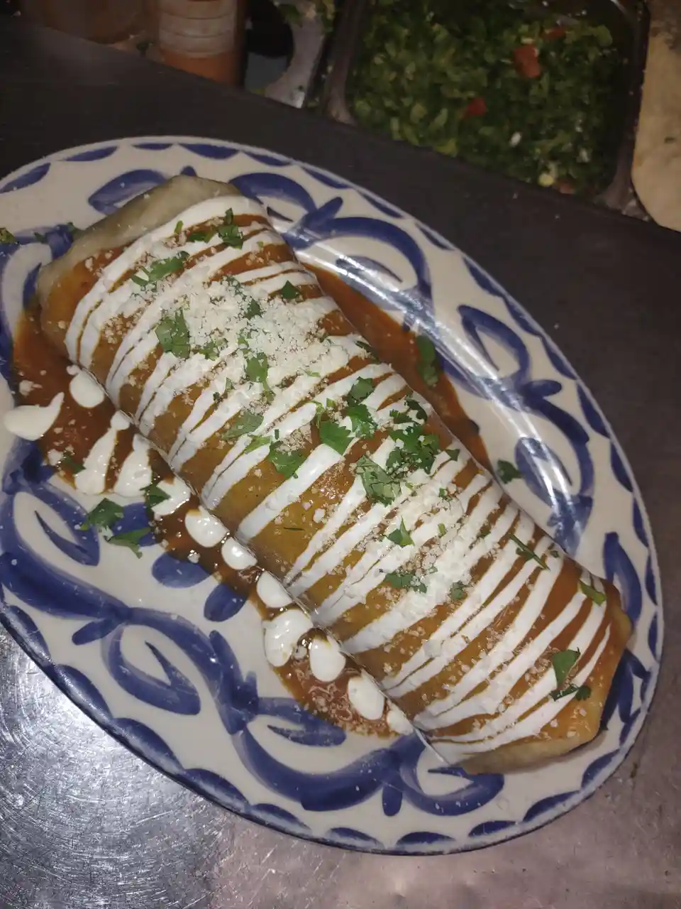 A close-up photo shows a single rolled enchilada on a blue and white patterned plate, covered in brown sauce, white cream…