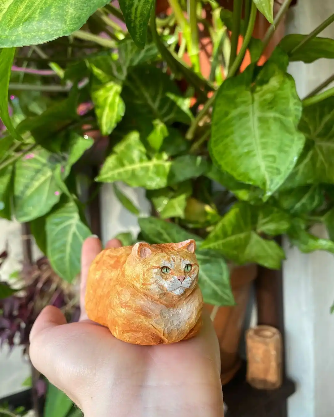 A hand holds a small, realistic sculpture of an orange tabby cat, crafted to look like a croissant, against a backdrop of…