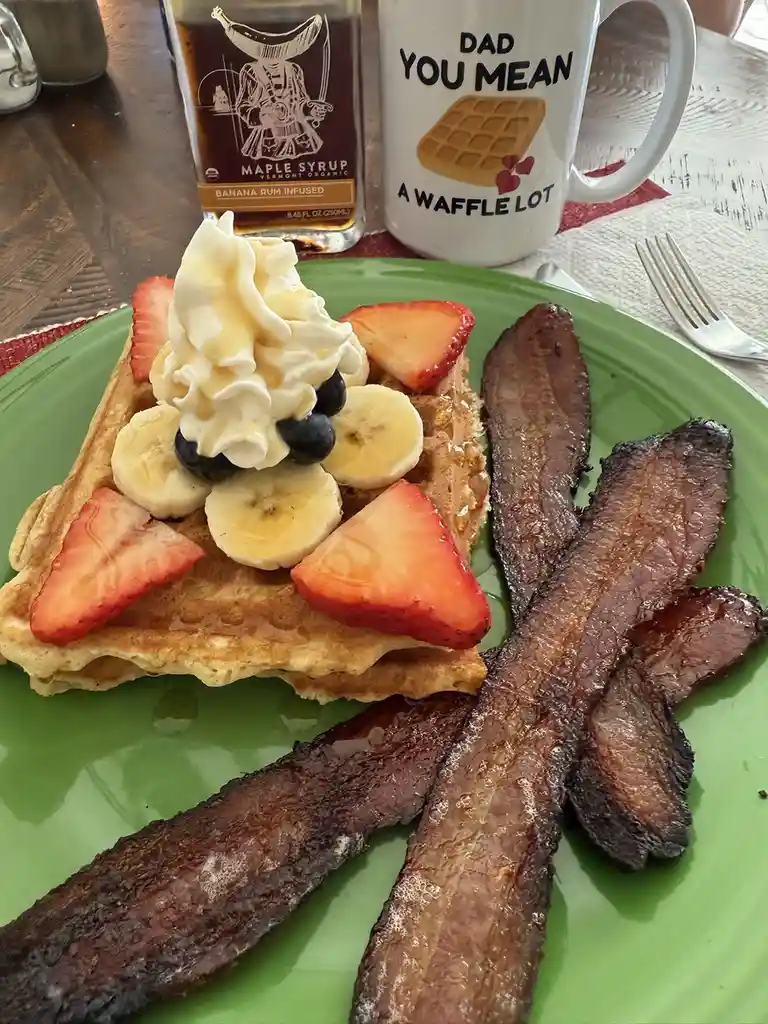 A green plate holds a waffle topped with whipped cream, sliced bananas, strawberries, and blueberries, alongside several…