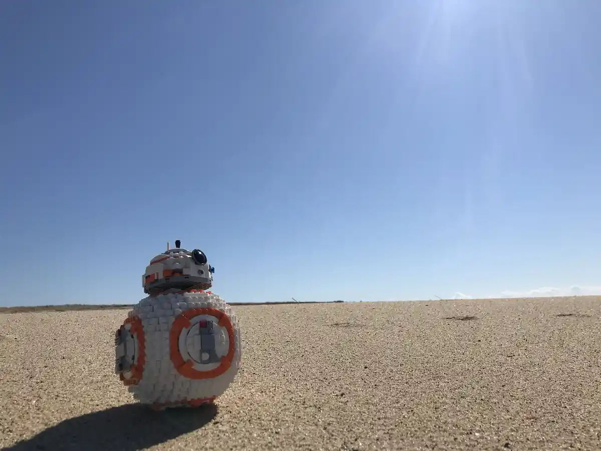A LEGO model of the Star Wars droid BB-8 stands alone on a vast, sandy beach under a bright blue sky with the sun shining…