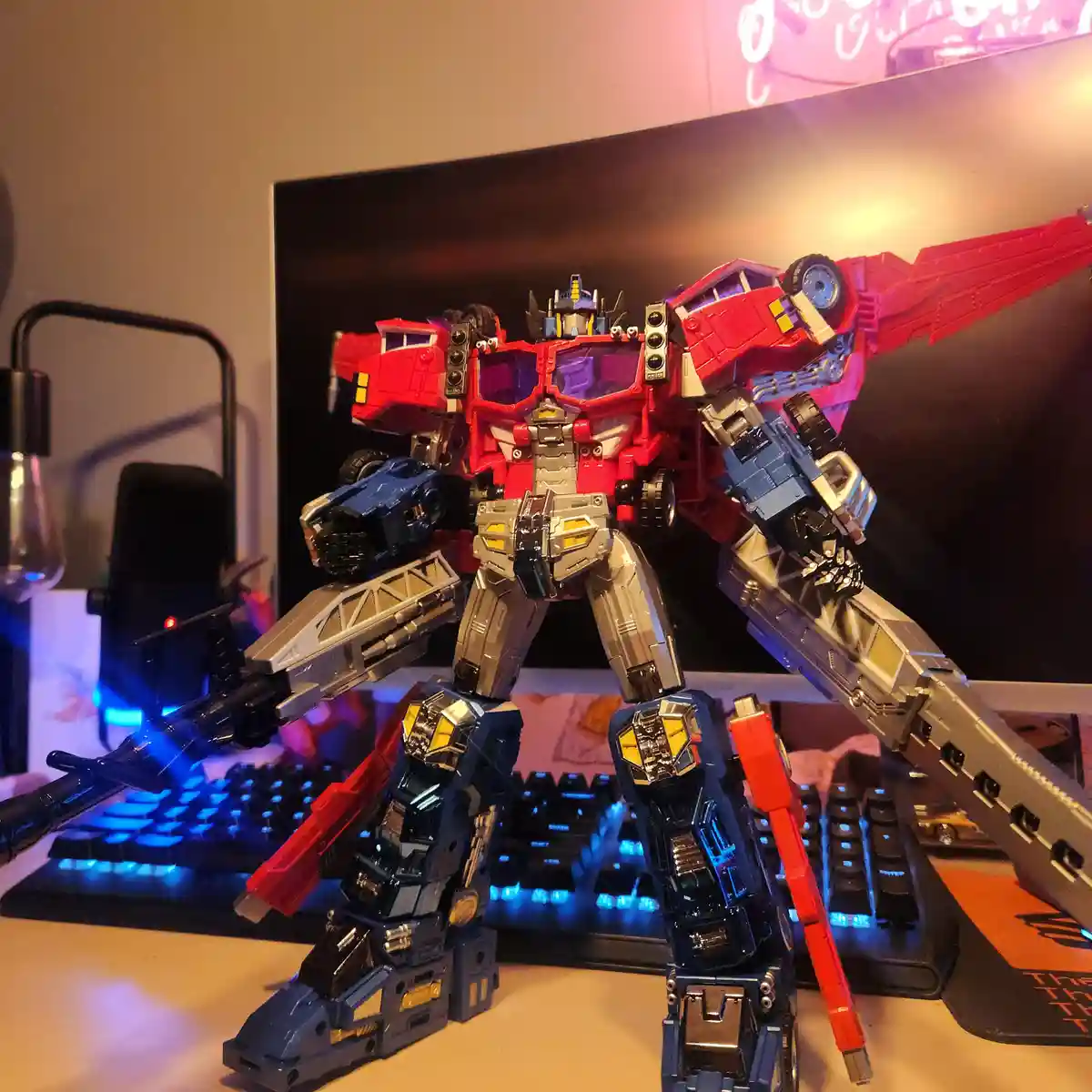 A large, red and blue Optimus Prime action figure stands on a desk in front of a computer monitor, with a glowing blue…