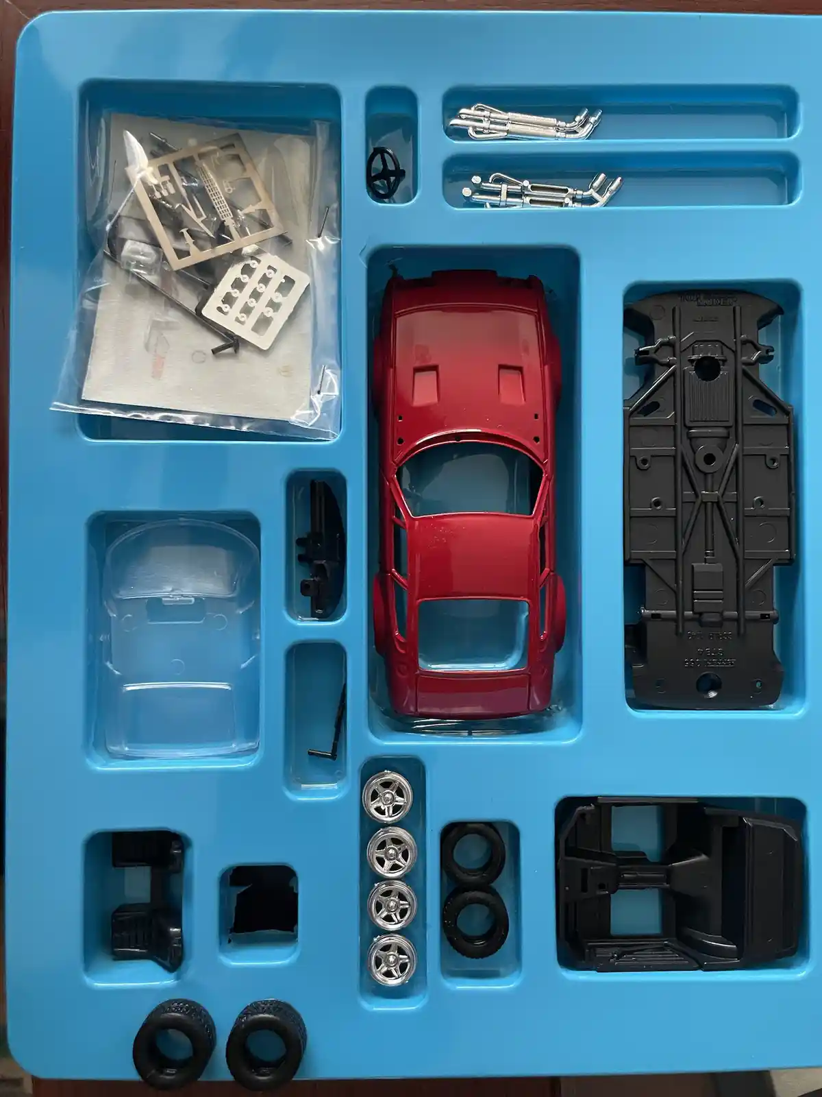 A top-down view of a blue plastic tray containing all the unassembled parts of a model car kit, including a red body shell…