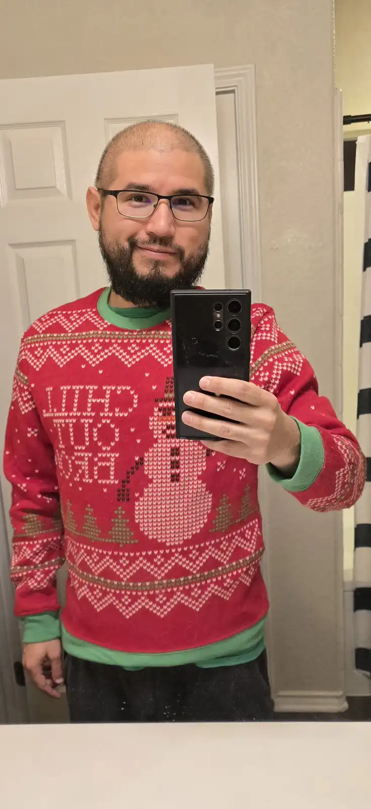 A man with glasses and a beard takes a mirror selfie wearing a red holiday sweater with a snowman and trees pattern. The…
