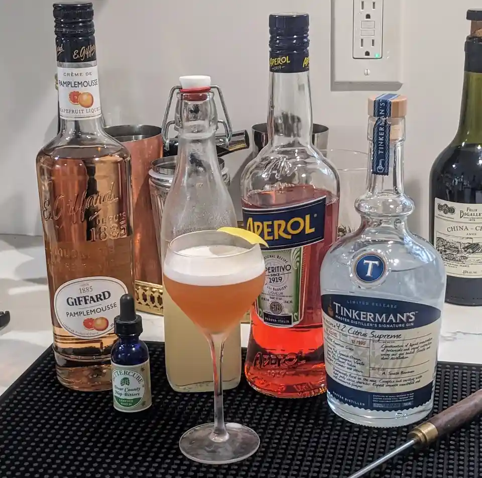 a cocktail in a coupe glass with a lemon twist sits on a bar mat surrounded by bottles of giffard pamplemousse liqueur…