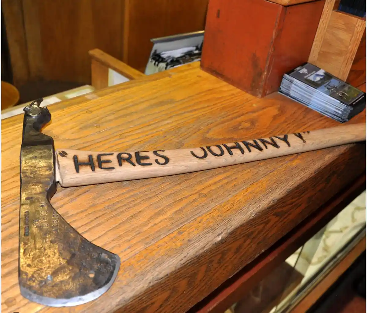 An old, rusty axe with a wooden handle rests on a wooden counter. The handle has the phrase 'HERES JOHNNY!' carved into it…