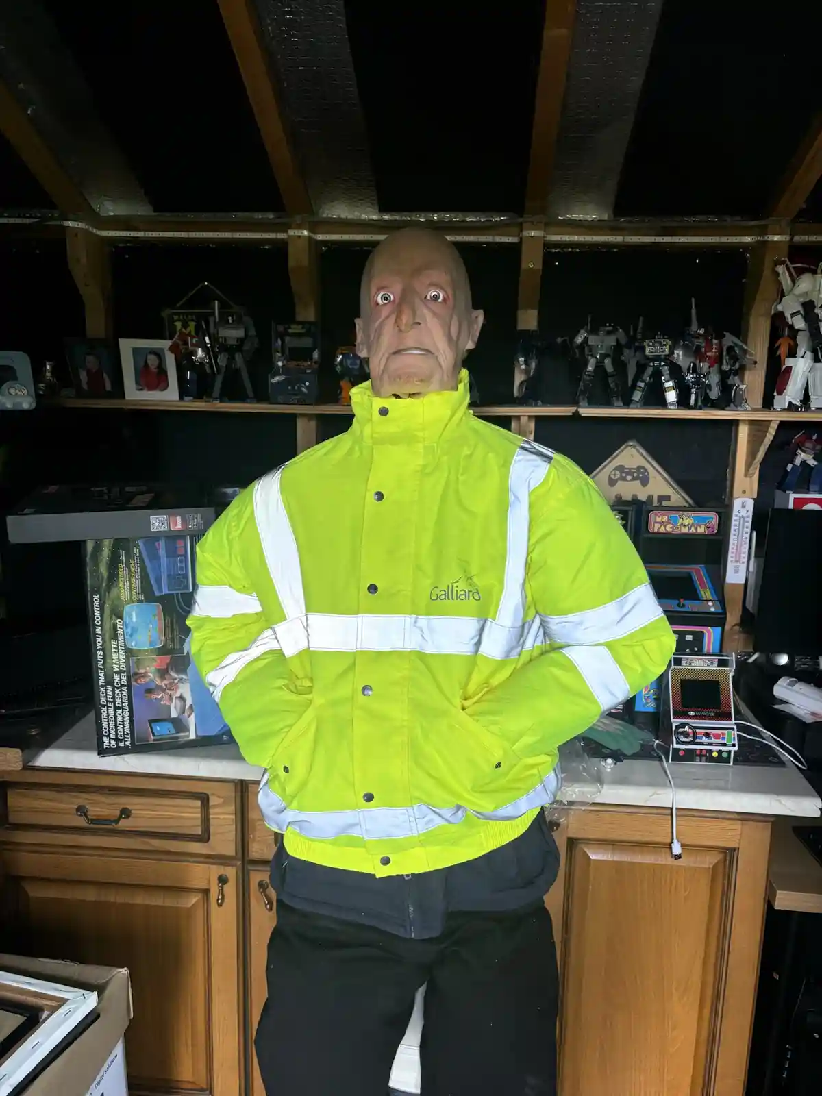A mannequin with a bald, wrinkled, and wide-eyed face stands in a room filled with retro gaming memorabilia, wearing a…