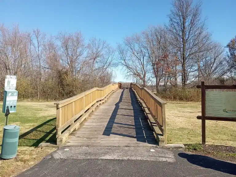 A wooden boardwalk bridge stretches into the distance under a clear blue sky, flanked by bare trees and grassy areas. On the…